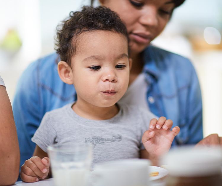 Niño pequeño sentado en la mesa sosteniendo una taza y mamá sentada detrás de él