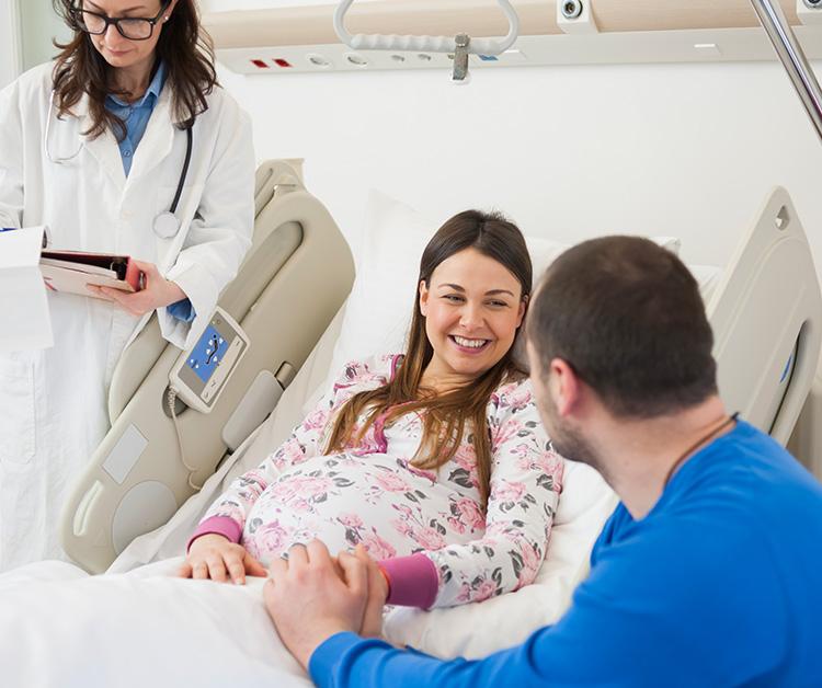 Pregnant woman laying in a hospital bed smiling as her spouse holds her hand with the doctor in the background