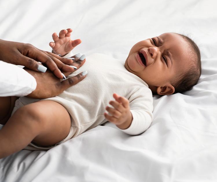 A baby lying on a white bed, crying, while an adult gently places their hands on the baby's stomach for comfort.
